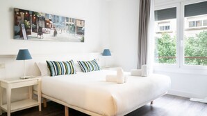 A neatly arranged hotel room with a large bed, two bedside tables, a painting on the wall, and a window with a view of buildings and trees. - Casa Cosi - Casanova 2 (Barcelona)