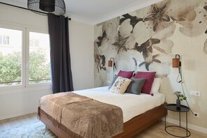 Un dormitorio con cama, ventana con cortinas, pared con papel tapiz floral y una pequeña mesa con un florero.