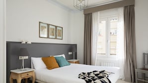 A neatly arranged bedroom with a large bed, bedside tables, a window with curtains, and framed pictures on the wall. - Casa Cosi - Casanova 4 (Barcelona)