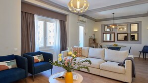 Ein Wohnzimmer mit einer Couch, zwei Sesseln, einem Couchtisch und einem Esstisch mit Stühlen. Es gibt ein großes Fenster mit Vorhängen, eine Chandelier und gerahmte Bilder an der Wand.