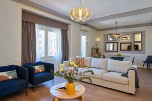 A living room with a sofa, two armchairs, a coffee table, and a dining table with chairs. There is a large window with curtains, a chandelier, and framed pictures on the wall. - Casa Cosi - Casanova 5 (Barcelona)