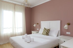A neatly made bed with white linens, a pillow with a leaf pattern, and a pair of towels on the bed. There are two bedside tables with lamps, and a window with sheer curtains allowing natural light. - Casa Cosi - Casanova 6 (Barcelona)