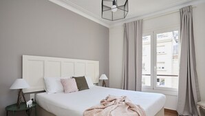A neatly made bed with a white headboard, two bedside lamps, and a window with curtains. - Casa Cosi - Casanova 7 (Barcelona)