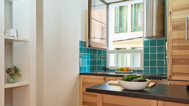 Una cucina con mobili in legno, un piano di lavoro nero, una finestra con persiane verdi e una ciotola di verdure.