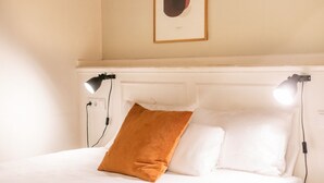Ein ordentlich bezogenes Bett mit weißen Leinen und einem einzelnen braunen Kopfkissen, zwei Nachttischlampen und ein gerahmtes abstraktes Kunstwerk an der Wand.