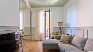 A living room with a grey sofa, a television on a stand, a floor lamp, and a view of the outside balcony.