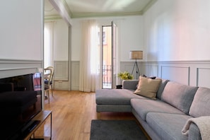 Un salón con un sofá gris, un televisor sobre un mueble bajo, una lámpara de pie y vistas al balcón exterior.