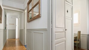 A hallway with white wainscoting, a wooden-framed mirror, and a chair in a room with a tiled floor.