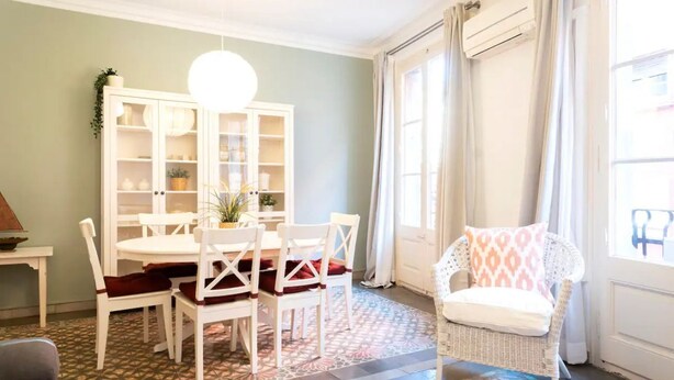 Un comedor con una mesa y sillas blancas, un armario blanco con puertas de cristal y una silla de mimbre con cojín.