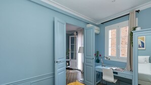 A room with a bed, a desk, and a chair. There is a window with curtains and a plant on the desk.