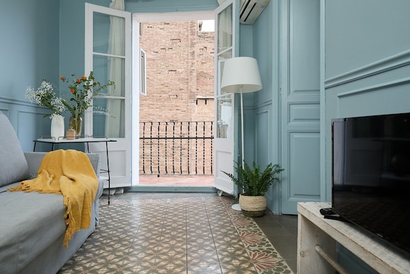 Una sala de estar con un sofá gris, una manta amarilla, una mesita con un jarrón de flores, una planta en maceta y un televisor sobre un mueble de madera.