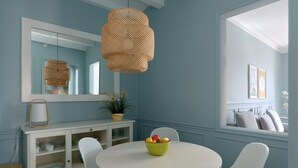 A dining area with a round table, white chairs, a bowl of fruit, a hanging woven light fixture, a white cabinet, and a large mirror.