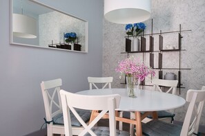 Un coin repas avec une table ronde, des chaises blanches, un vase de fleurs, une étagère fixée au mur et un grand miroir.