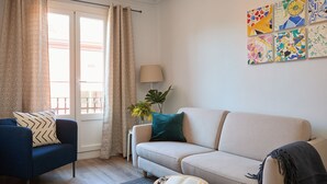 Ein Wohnzimmer mit blauem Sessel, beiger Couch, rundem Couchtisch und offenem Buch sowie einer Lampe.