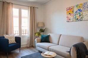 Un salon avec un fauteuil bleu, un canapé beige, une table basse ronde sur laquelle est posé un livre ouvert, et une lampe.
