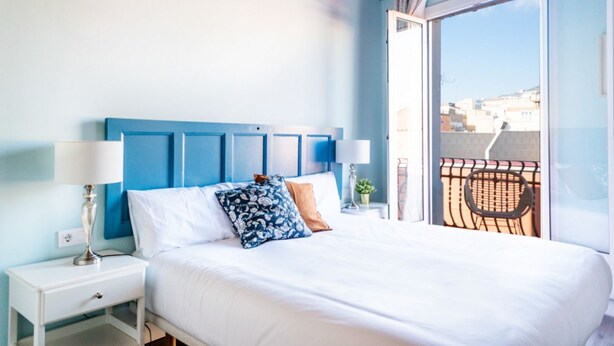 Une chambre à coucher comprenant un lit, deux tables de chevet, une fenêtre donnant sur des immeubles et un balcon avec une chaise.