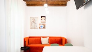 Un salon moderne avec un canapé blanc, une petite table noire et une table à manger blanche avec des chaises vertes. Des tableaux encadrés ornent le mur et un téléviseur est fixé au-dessus du canapé.