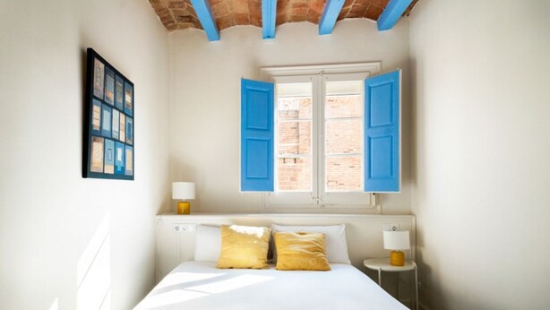 Un dormitorio con cama, dos lámparas de noche, una ventana con contraventanas azules y un cuadro en la pared.