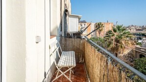 Un balcón con una silla blanca y una mesa pequeña, con vistas a un paisaje urbano con edificios y palmeras.
