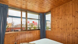 Una habitación con paredes y techo de madera, una cama con colchón blanco y manta marrón, y un ventanal con cortinas azules.