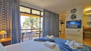 Habitación de hotel con cama, escritorio, silla, televisor y balcón con vista a palmeras.