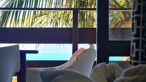 Una habitación con vista al océano, una cama con almohada blanca y naranja, y una ventana que da a palmeras.