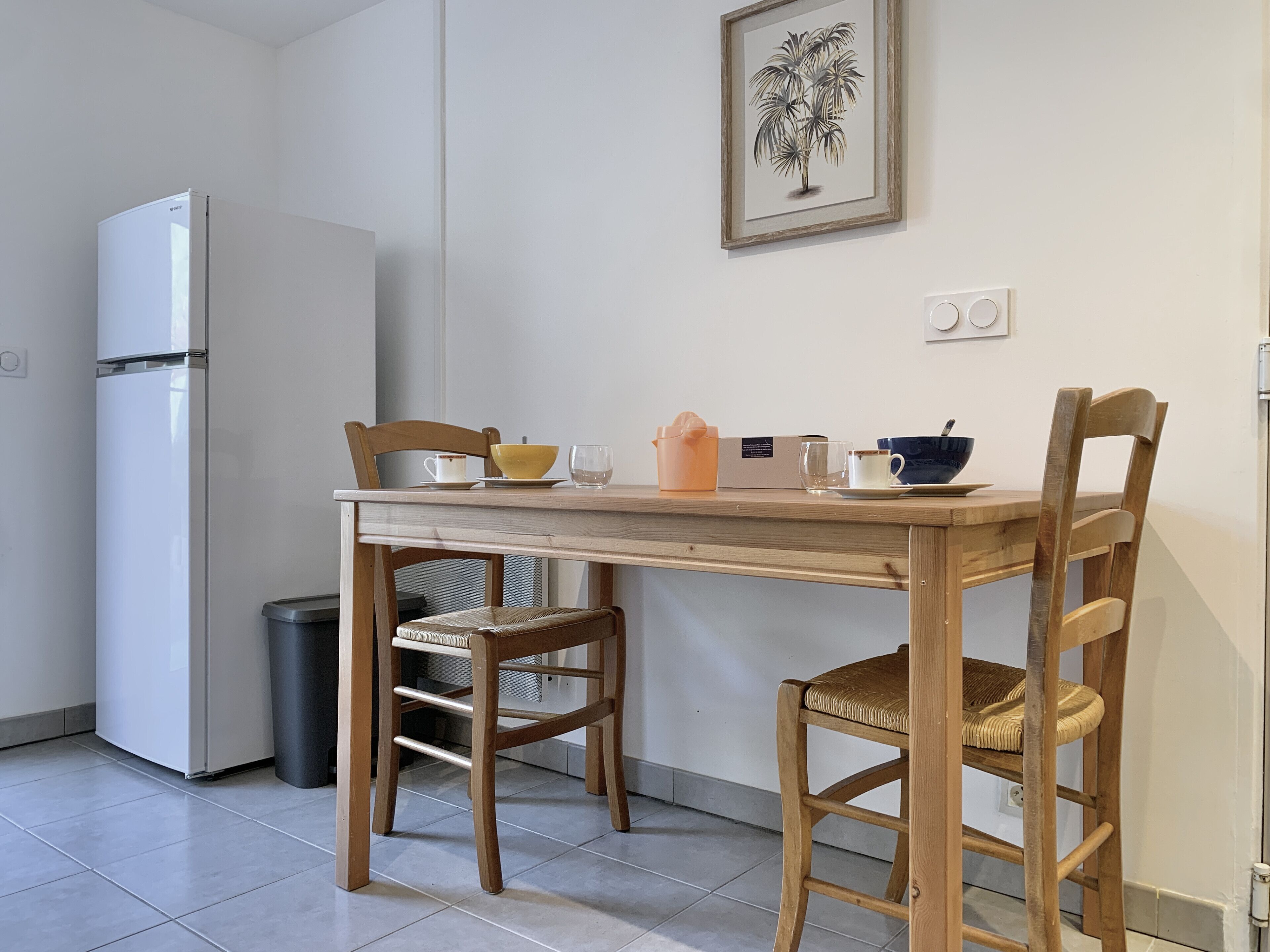 A compact kitchen with a white refrigerator, a wooden dining table, two wicker chairs, and a framed botanical print on the wall.