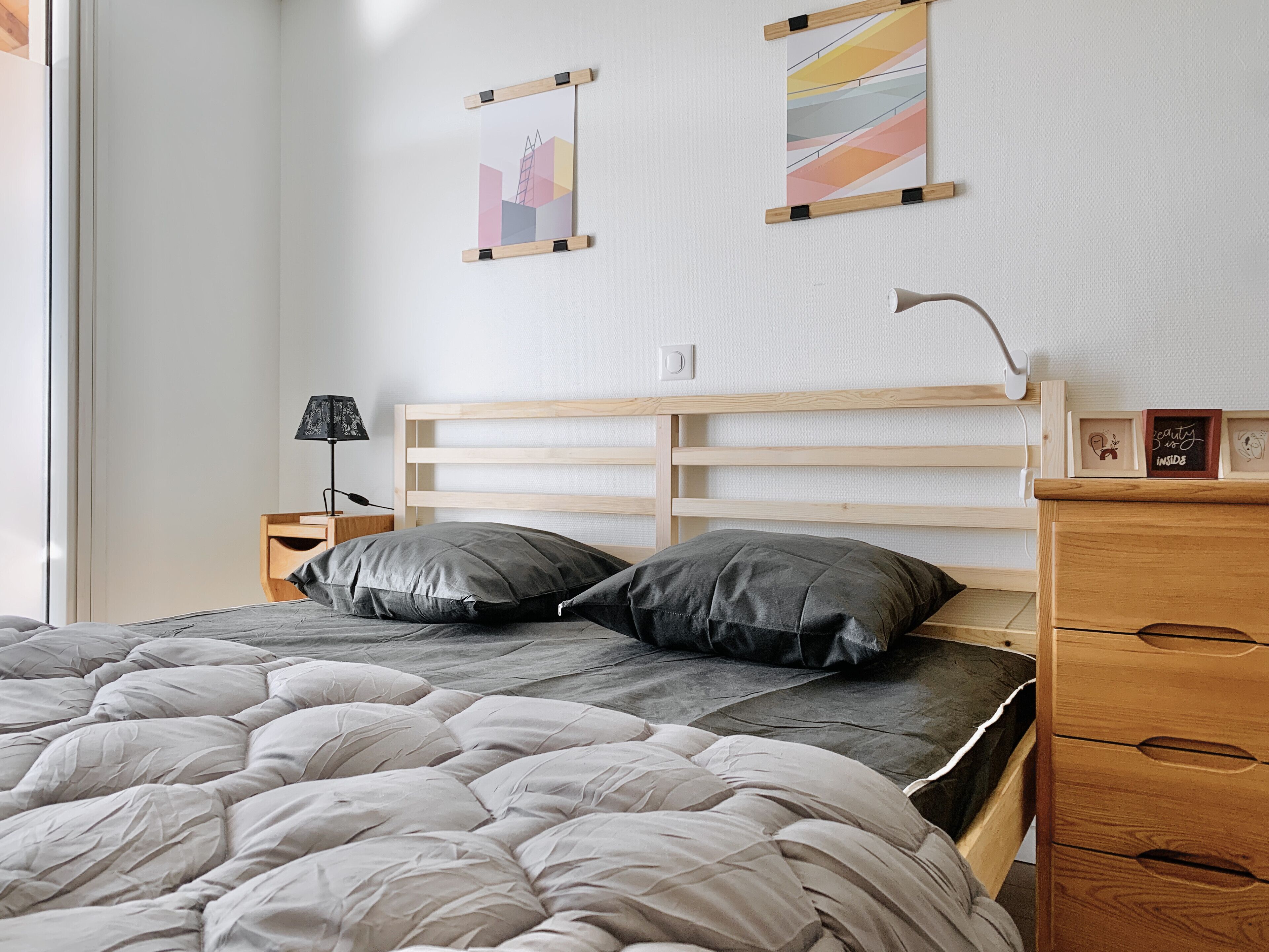Un dormitorio con cabecera de madera, cama con ropa de cama gris, mesita de noche con lámpara y dos obras de arte en la pared.