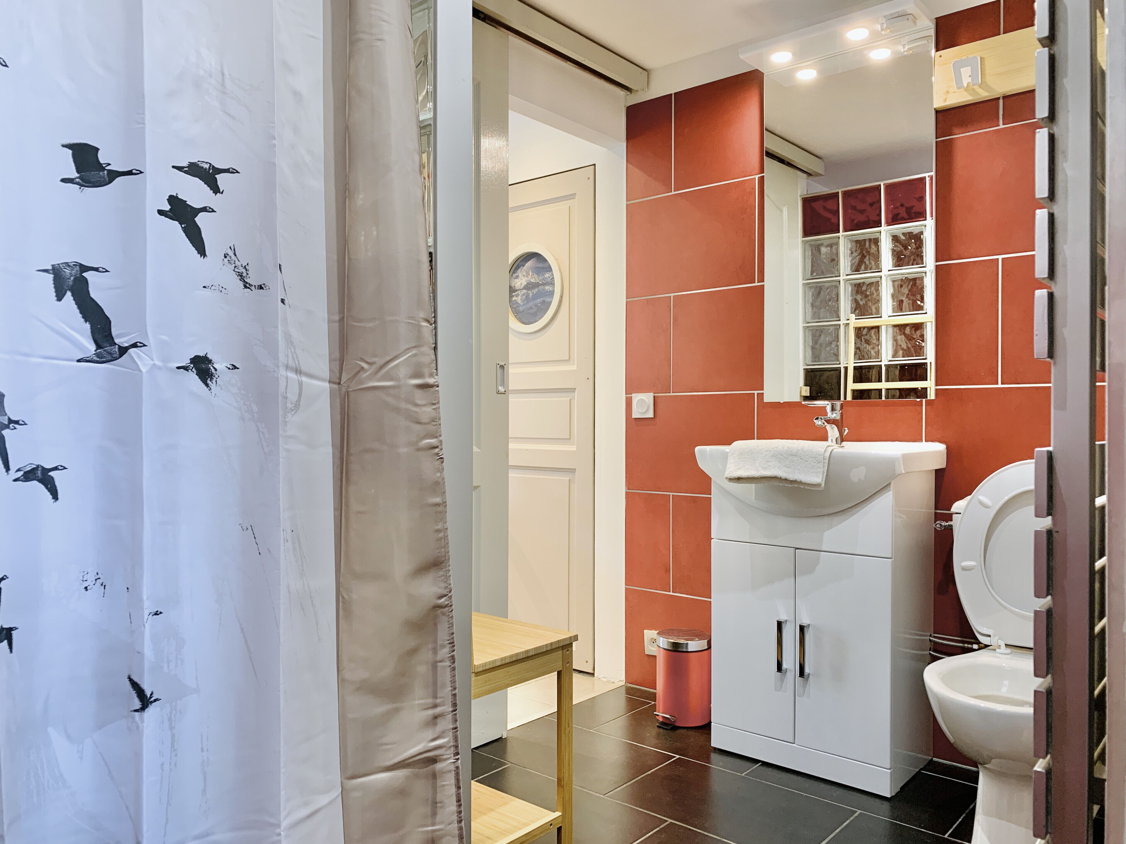 A bathroom with a white sink, red-tiled walls, a white toilet, and a small wooden stool.