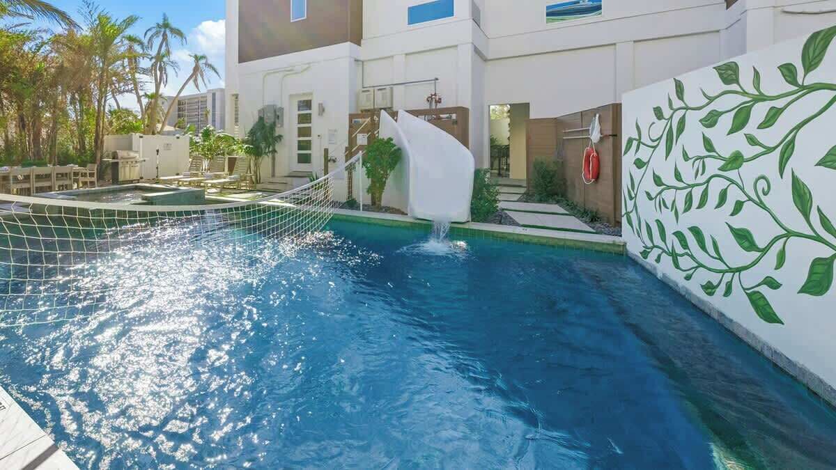 A swimming pool with a waterfall feature, surrounded by a modern building with a white exterior and green leaf patterns on the wall.