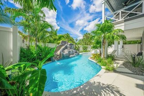 A swimming pool with a waterfall feature, surrounded by lush greenery and a modern building. - Resort Pool w/Slide - Walk to Beach & Village (Sarasota)