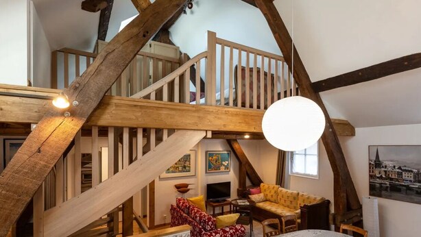 Ein Loft-Wohnbereich mit freiliegenden Holzbalken, einer Treppe, einer Couch, einem Fernseher und einem Gemälde an der Wand.