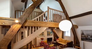 A loft-style living area with exposed wooden beams, a staircase, a sofa, a television, and a painting on the wall.