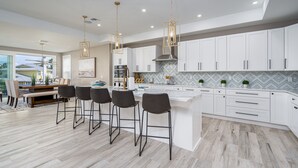 Una cocina moderna con armarios blancos, una isla grande, taburetes de bar, luces colgantes y vistas al exterior.