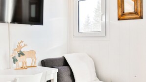 A modern room with a grey sofa, a white desk with a chair, a flat-screen TV mounted above the desk, a window with a view of trees, and a framed picture on the wall. - Immodreams - Le Vivace - Avoriaz (Avoriaz)