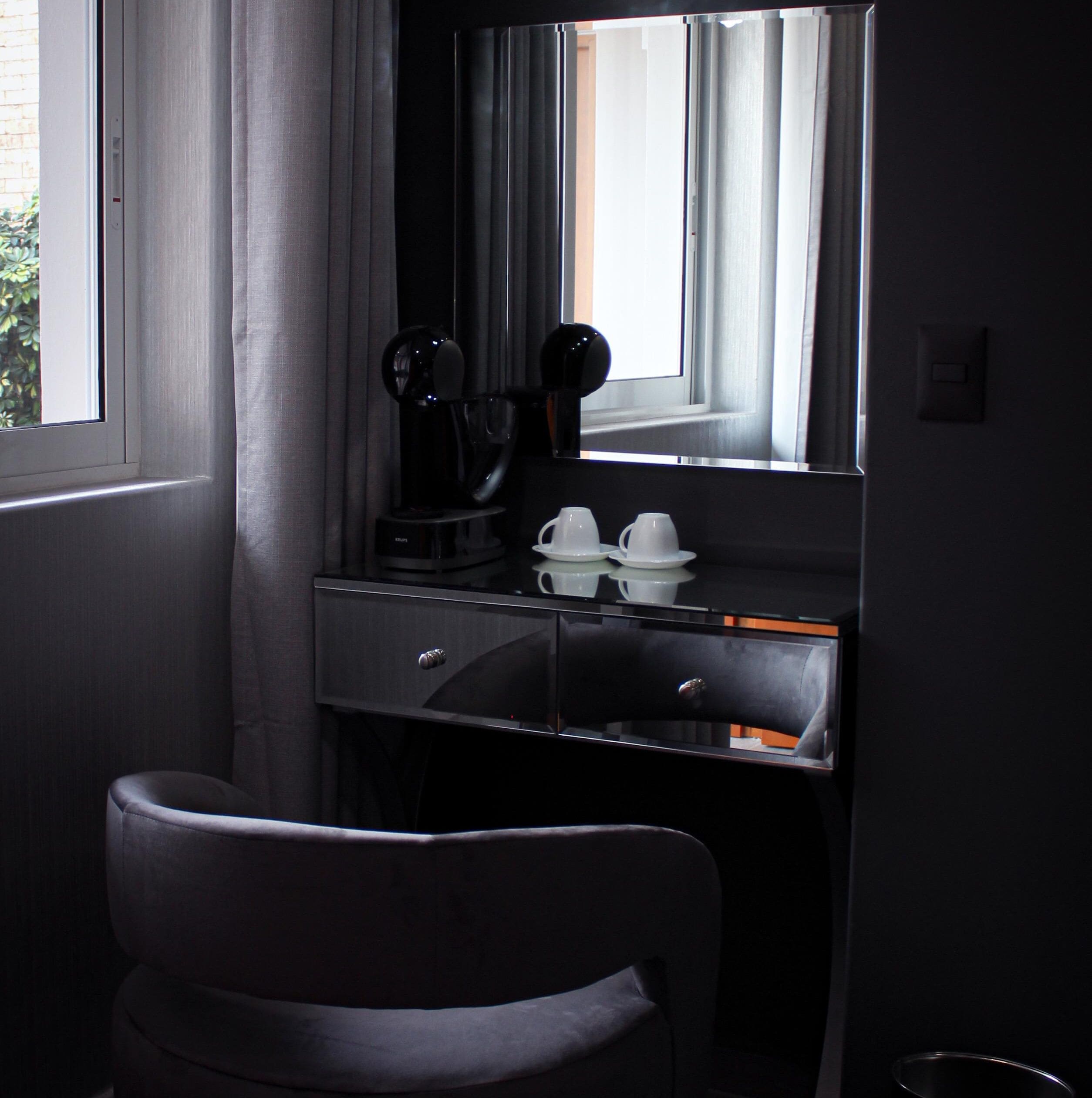 Una habitación de hotel pequeña con un escritorio de color oscuro, una cafetera y un espejo que refleja la ventana y las cortinas.