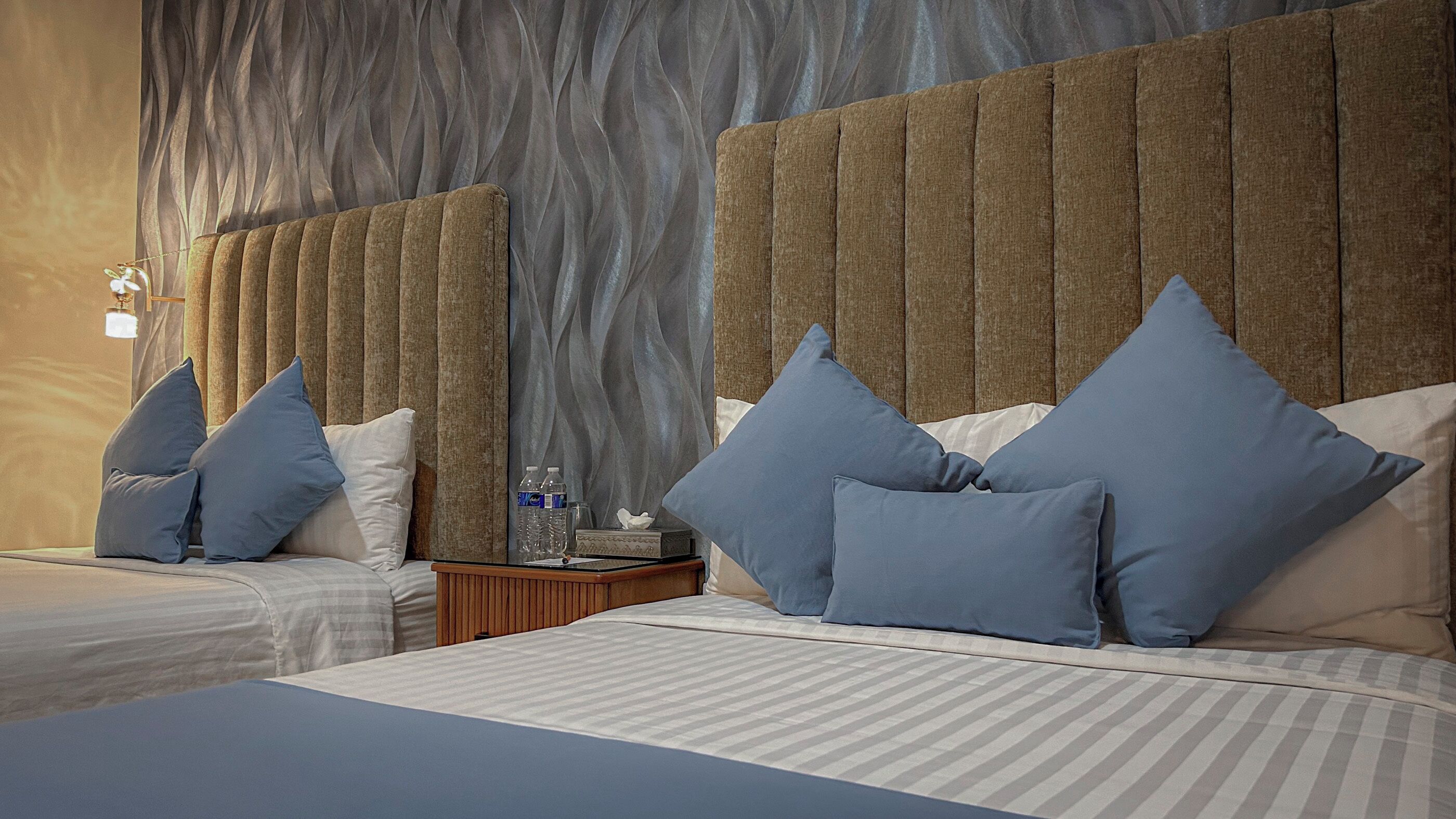 A hotel room with two beds, each with blue pillows and a striped bedspread. There is a wooden nightstand with a water bottle and a tissue box. The room has a textured wall and a wooden headboard.