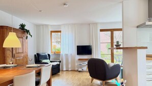 A modern living room with a dining area, a flat-screen TV, and a large window with curtains.