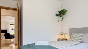 A bedroom with a bed, a hanging plant, a bedside table with a lamp, and a view of a living area with a chair and a TV.