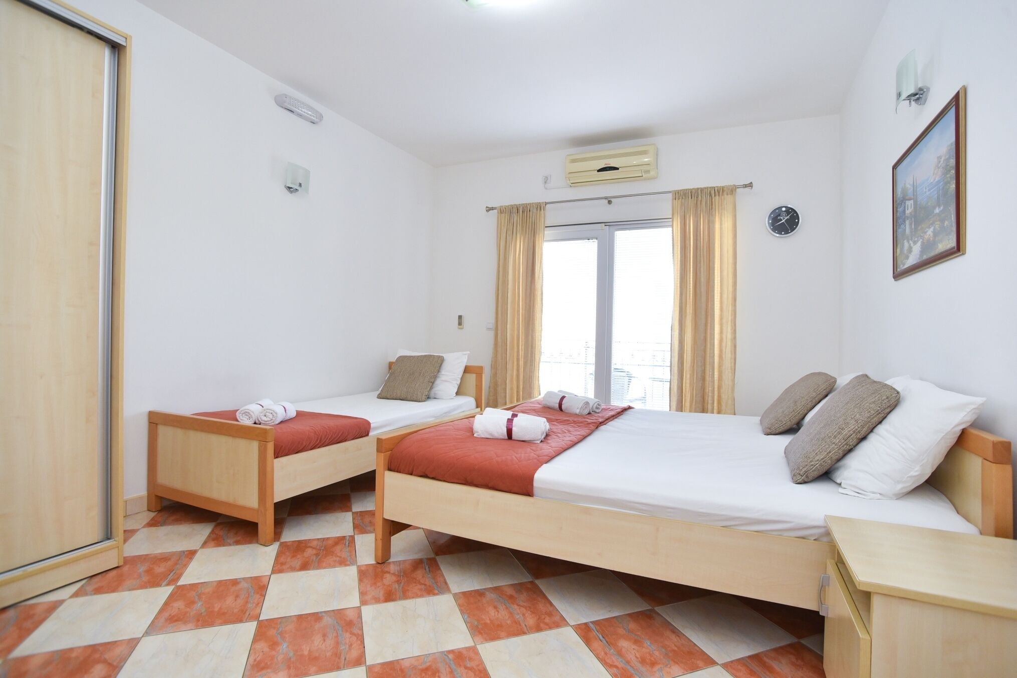 Une chambre d’hôtel avec deux lits en bois, une fenêtre avec des rideaux, une horloge et un tableau encadré au mur.
