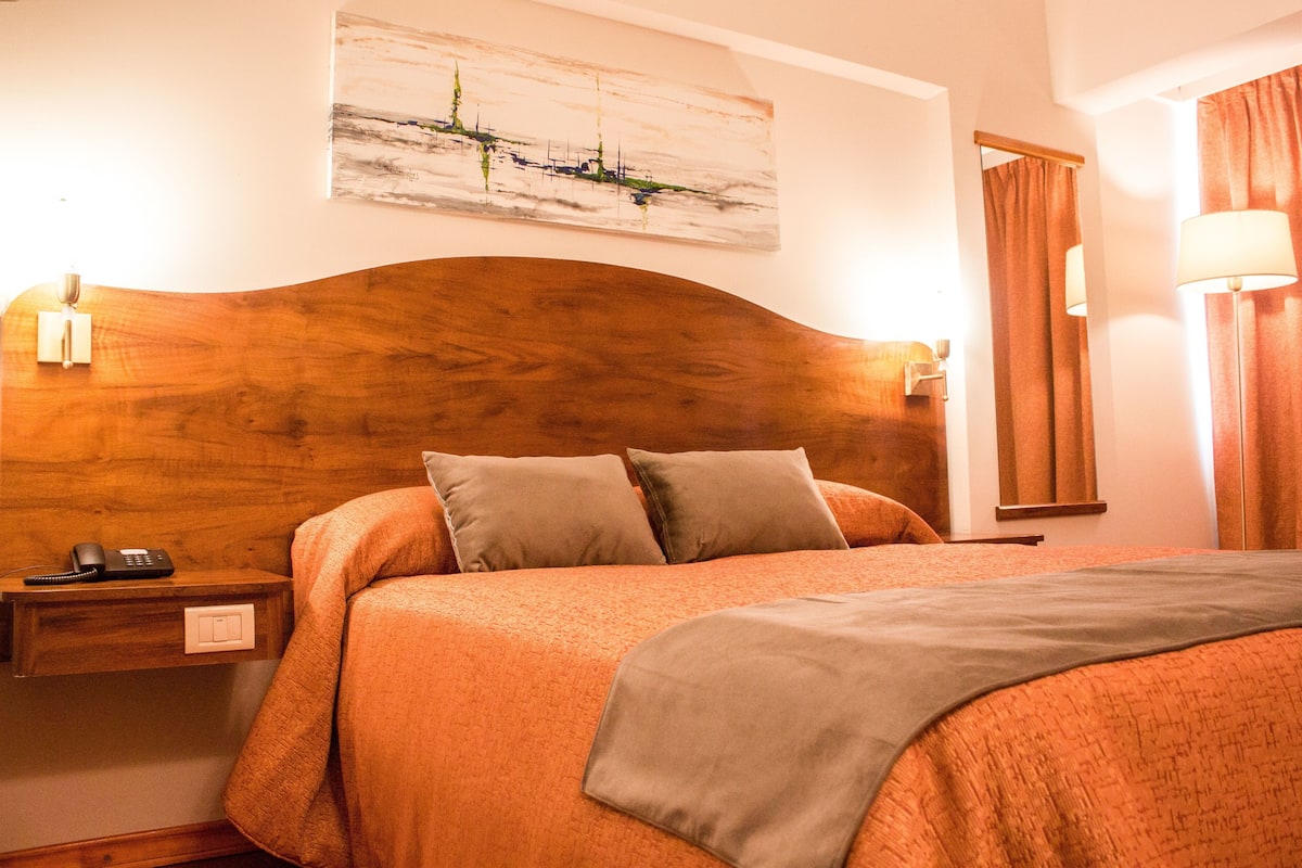 A hotel room with a wooden headboard, a bed with orange bedding, a nightstand with a phone, and a painting on the wall.