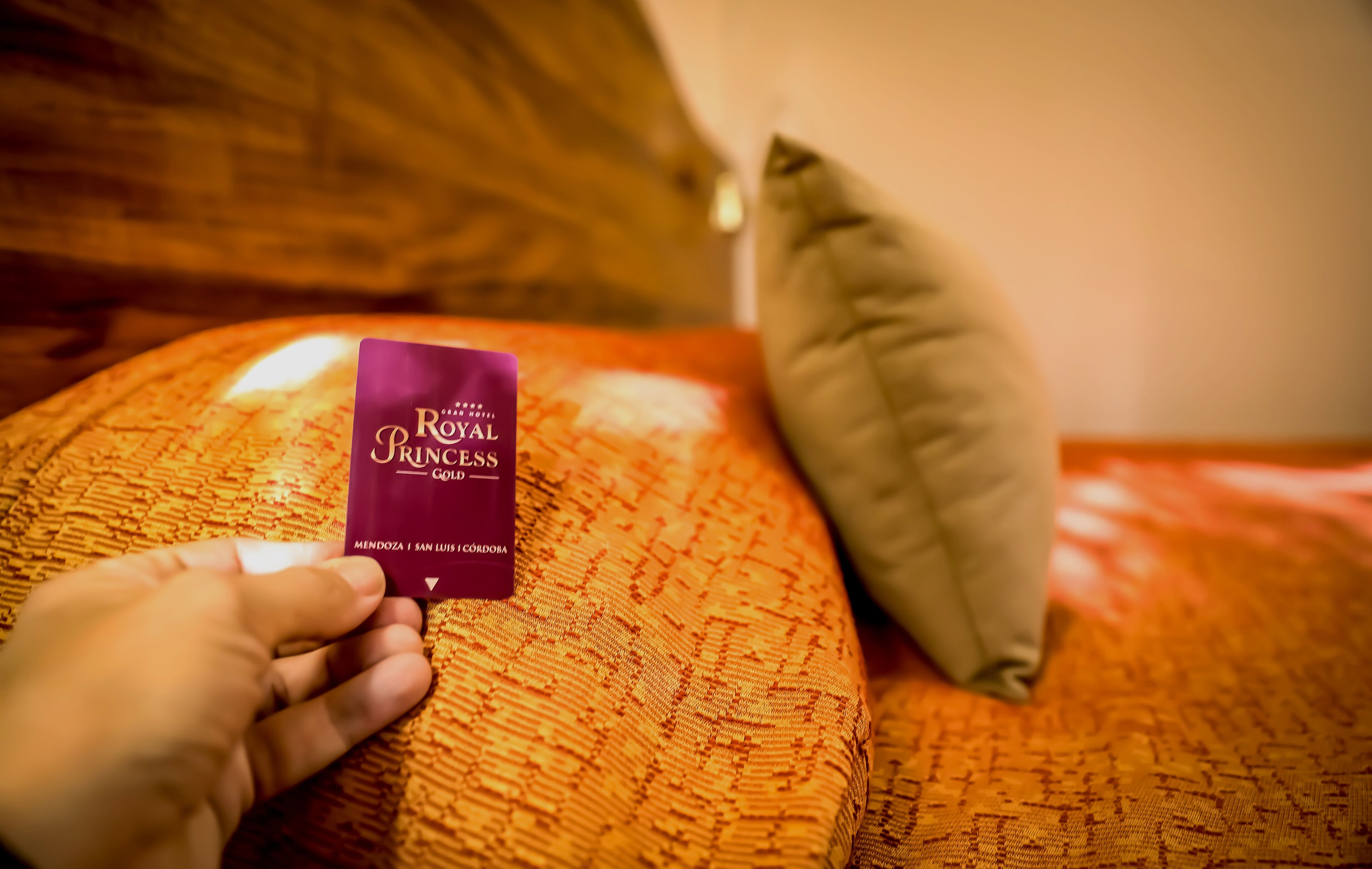 Una mano sostiene una tarjeta morada con la etiqueta 'Royal Princess' sobre una colcha anaranjada con textura y almohadas.