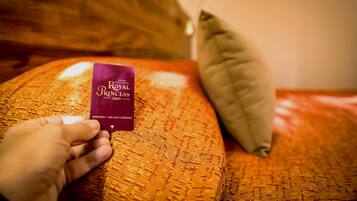 Una mano sostiene una tarjeta morada con la etiqueta 'Royal Princess' sobre una colcha anaranjada con textura y almohadas.