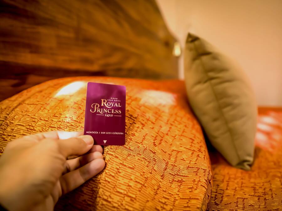 Una mano sostiene una tarjeta morada con la etiqueta 'Royal Princess' sobre una colcha anaranjada con textura y almohadas.