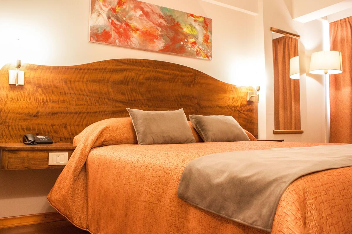 A hotel room with a wooden headboard, two pillows, a bedside table with a phone, and a painting on the wall.