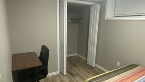 A small room with a wooden desk, a black office chair, a striped bed, and a closet with a door open.