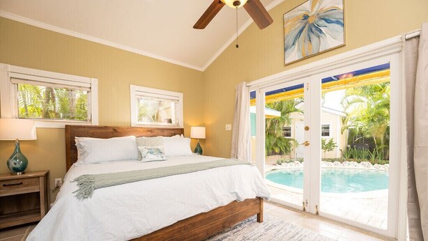 A bedroom with a wooden bed, two bedside tables with lamps, a large window, and a view of a swimming pool. - BE STILL- 4 Bedroom, 3 Bath with POOL & Guest Cottage (Key West)