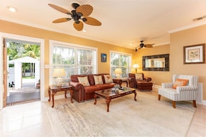 A spacious living room with a brown leather sofa, a striped armchair, and a coffee table. There are two ceiling fans, a large window with blinds, and a door leading to a covered outdoor area. - BE STILL- 4 Bedroom, 3 Bath with POOL & Guest Cottage (Key West)