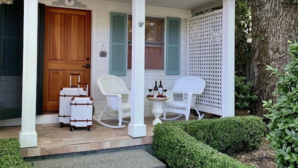 A house with a porch, a wooden door, and two rocking chairs. There are two suitcases and a table with bottles on the porch.