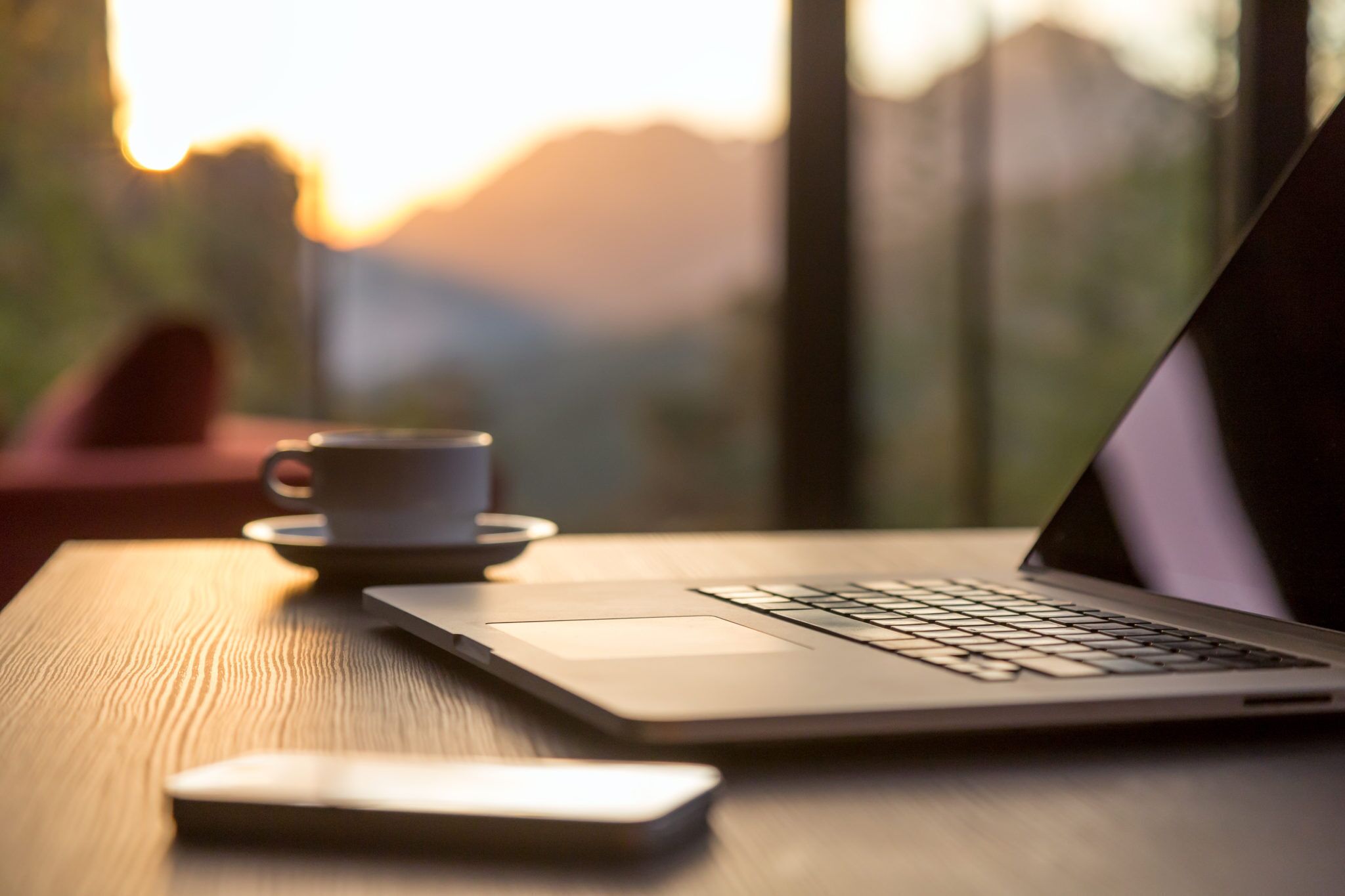 A laptop on a wooden table with a smartphone and a cup.